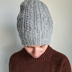 May include: A gray cable-knit beanie hat. The hat is made of a soft, textured yarn and features a ribbed brim. The beanie is a neutral color and is suitable for cold weather. The hat is a stylish accessory.