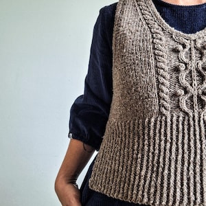 Op de afbeelding: Een bruin gebreid vest met een kabelpatroon aan de voorkant en geribbelde details. Het vest wordt gedragen over een donkerblauw shirt met lange mouwen. Het vest heeft een V-hals en een getailleerd silhouet, wat een klassieke, handgemaakte uitstraling geeft.