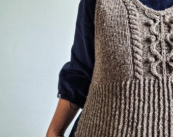 May include: A brown knitted vest with a cable knit design on the front and ribbed detailing. The vest is worn over a dark blue long-sleeved shirt. The vest has a V-neck and a fitted silhouette, showcasing a classic, handmade aesthetic.