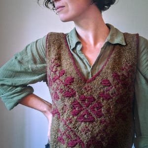 Può includere: Un gilet lavorato a maglia marrone e bordeaux con scollo a V. Il gilet presenta un motivo floreale ripetuto in bordeaux. La persona nell'immagine indossa una camicia verde abbottonata e una gonna blu scuro.