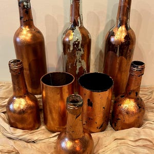 Può includere: Una collezione di bottiglie di vetro e contenitori cilindrici riutilizzati, dipinti con una finitura metallica color rame. Le bottiglie variano per dimensioni e forma, alcune con resti del colore originale del vetro. La disposizione suggerisce un tema decorativo o di riciclo.