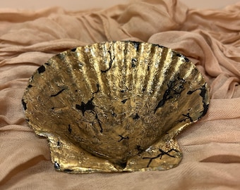 Puede incluir: Un plato decorativo en forma de concha con acabado de pan de oro y detalles negros. El plato descansa sobre una tela color melocotón, creando un contraste entre las texturas y los colores. Se ven las estrías de la concha.