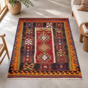 May include: A rectangular area rug featuring a central red panel with geometric diamond motifs in white, orange, and green. The rug is bordered by black, orange, and green bands with zigzag patterns and finished with white fringe on both ends.