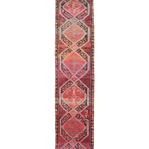 Puede incluir: Una alfombra larga y rectangular con un patrón geométrico. La alfombra presenta un diseño central de formas de diamante repetidas en tonos de rojo, naranja y rosa, delineadas en negro. Los bordes de la alfombra están bordeados con un ribete más oscuro y estampado.