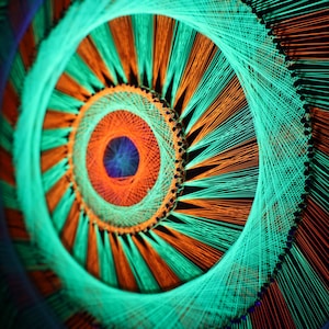 Könnte beinhalten: Abstrakte Kunst mit einem Spiralmuster aus konzentrischen Kreisen. Das Kunstwerk verwendet orange, türkis und blaue Fäden vor einem schwarzen Hintergrund. Die Fäden sind sorgfältig angeordnet und erzeugen ein visuell auffälliges und komplexes Muster.