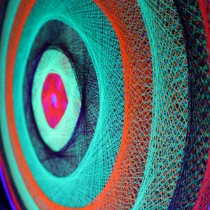 Op de afbeelding: Een close-up van een kleurrijk draadkunstwerk. Concentrische cirkels van levendige kleuren, waaronder oranje, blauwgroen, zwart en paars, creëren een fascinerend spiraalontwerp. Het middelpunt is felroze. Het kunstwerk staat tegen een donkere achtergrond.