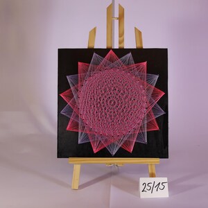Può includere: Un'opera d'arte con filo su un piccolo cavalletto di legno. L'opera presenta un design geometrico con fili rosa e bianchi su sfondo nero. Il pezzo è etichettato "25/15".
