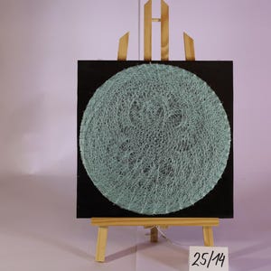 Può includere: Una tela quadrata nera con un design a cerchio in filo blu chiaro, esposta su un piccolo cavalletto di legno. La string art ha un aspetto strutturato. Il numero "25/14" è visibile su una piccola etichetta bianca.