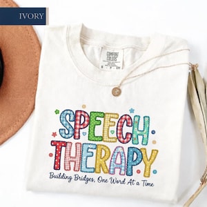 Könnte beinhalten: Ein elfenbeinfarbenes Kurzarm-T-Shirt mit dem Schriftzug "SPEECH THERAPY" in großen, bunten Buchstaben mit verschiedenen Mustern, die an Patchwork erinnern. Darunter steht "Building Bridges, One Word At a Time" in dunkelblauer Schrift.
