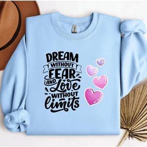 Könnte beinhalten: Hellblaues Sweatshirt mit dem Text "DREAM WITHOUT FEAR AND LOVE WITHOUT LIMITS" in Schwarz. Rosa und lila Herzen befinden sich auf der rechten Seite. Ein brauner Hut befindet sich in der oberen linken Ecke.