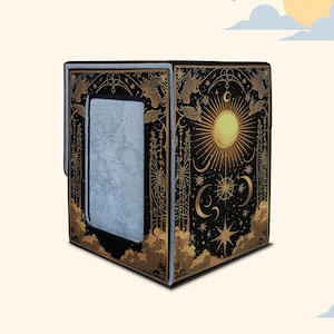 May include: A black deck box featuring gold foil celestial illustrations of a sun, moon, and stars. The front panel has a rectangular cutout revealing a grey interior lining. The design includes intricate line art of clouds and birds.