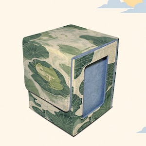 May include: A square deck box featuring a green frog resting on a large lily pad surrounded by white lotus flowers and green leaves. The exterior has a textured fabric finish, and the front panel reveals a solid light blue interior lining.