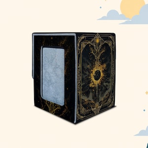 Puede incluir: Una caja para barajas negra con filigrana dorada y arte celestial. El panel frontal muestra un motivo central de sol y luna rodeado de bordes decorativos. El panel lateral presenta un área rectangular gris con textura.