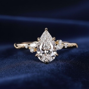 Könnte beinhalten: Ein goldener Verlobungsring mit einem birnenförmigen Diamanten. Der Ring hat kleinere Akzentsteine und ein strukturiertes Band. Der Diamant funkelt vor einem dunkelblauen Hintergrund und zeigt das aufwendige Design des Rings.