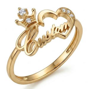 Puede incluir: Un anillo de tono dorado que presenta la palabra Custom en letra cursiva, acentuado por un motivo de corona a la izquierda y una forma de corazón a la derecha. La corona y el corazón están adornados con pequeñas piedras redondas transparentes.