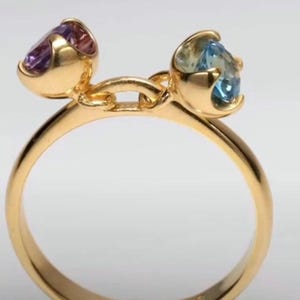 Könnte beinhalten: Ein Goldring mit zwei separaten Fassungen, die durch einen kleinen Goldsteg verbunden sind. Eine Fassung enthält einen runden, facettierten violetten Edelstein, die andere einen runden, facettierten hellblauen Edelstein, beide in Goldkrappen gefasst.