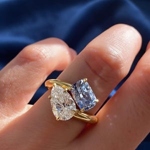 Peut inclure: Une bague en or ornée de deux pierres précieuses : un diamant clair en forme de poire et un diamant bleu clair rectangulaire. La bague a un anneau en or poli. Les pierres sont serties de griffes en or.
