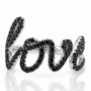 Puede incluir: Un anillo de tono plateado que presenta la palabra love escrita en letra cursiva. La superficie de las letras está incrustada con numerosas piedras negras pequeñas y redondas que cubren toda la parte frontal del diseño del anillo.