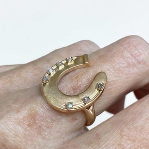 Op de afbeelding: Een goudkleurige hoefijzervormige ring met kleine, vierkant geslepen edelstenen. De ring wordt aan een vinger gedragen en toont zijn unieke ontwerp. De hoefijzervorm is een klassiek symbool van geluk.