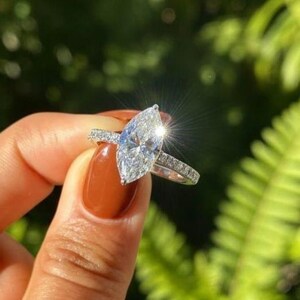 Könnte beinhalten: Ein silberfarbener Ring mit einem großen, marquise-geschliffenen Mittelstein und einer Ringschiene, die mit kleinen, klaren, rund geschliffenen Steinen besetzt ist. Der Mittelstein reflektiert helles Licht und erzeugt einen Sterneneffekt.