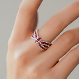 May include: A silver-toned ring featuring a bow-shaped design encrusted with small, vibrant red gemstones and tiny clear white stones. The band is constructed of multiple intersecting rows that form a knot-like center, creating a detailed piece.
