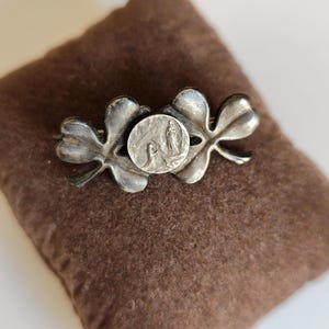 May include: A silver-toned metal brooch featuring two connected four-leaf clover shapes. Centered between the clover leaves is a small, circular medallion embossed with a detailed relief depicting two figures in a kneeling or standing pose.