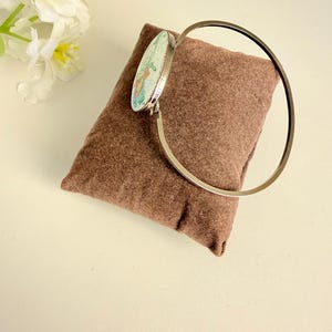 Pode incluir: Uma pulseira de metal prateado com um pingente circular que apresenta um fundo verde claro e uma ilustração floral colorida. A pulseira é exibida sobre uma almofada de tecido retangular castanha ao lado de flores artificiais brancas.