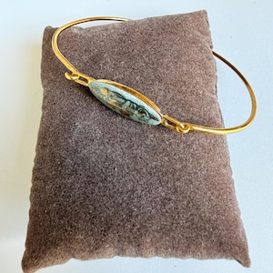 Peut inclure: Bracelet jonc doré avec une pierre ovale au centre. La pierre présente des nuances de bleu, de gris et d'or. Le bracelet est posé sur une surface marron texturée. Un bijou délicat.
