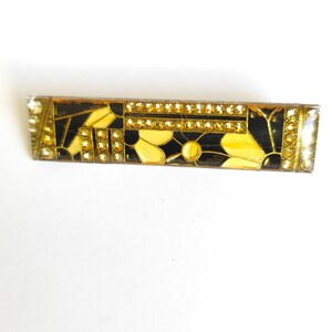 Peut inclure: Une barrette à cheveux rectangulaire ornée d'un motif floral noir et jaune avec des cristaux dorés incrustés. L'accessoire possède un cadre métallique et est décoré de rangées de petites pierres jaunes scintillantes sur les bords et au centre.