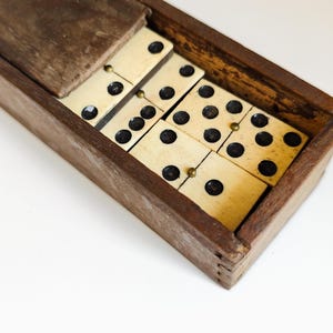Peut inclure: Une boîte rectangulaire en bois avec un couvercle coulissant contenant un jeu de dominos blanc cassé. Chaque domino présente des points noirs et une petite tige métallique couleur laiton au centre. La boîte présente des assemblages visibles.