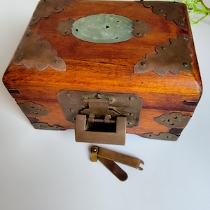 May include: A rectangular wooden box featuring brass corner plates, a central oval jade inlay on the lid, and a brass latch mechanism on the front. A separate brass key component with two flat prongs lies on the surface in front of the box.