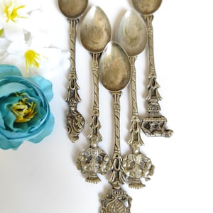 Può includere: Sei cucchiai antichi color argento con manici ornati e finiture decorative. I cucchiai hanno coppette a forma di goccia e disegni dettagliati sui manici, tra cui figure e stemmi. I cucchiai sono disposti su una superficie bianca.