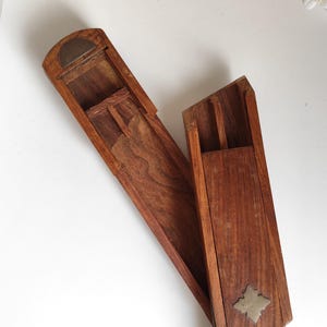 May include: A long, rectangular wooden box with a sliding lid, featuring a visible wood grain pattern and a small, metallic, star-shaped inlay on the exterior. The box is open, revealing a hollow interior compartment with a curved metal edge.