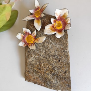 Peut inclure: Trois ornements floraux en métal fabriqués à la main, dotés de pétales blancs avec des centres rouge foncé et des étamines jaune vif. Les fleurs ont des bords sombres et définis, disposées sur une pierre naturelle grise et brune.