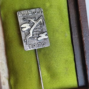 Peut inclure: Épingle argentée avec le texte "A.I.E.J.I." et l'année "1970" sur une plaque rectangulaire. L'épingle représente une image stylisée d'une gazelle sautant au-dessus d'un globe. L'épingle est posée sur un fond de velours vert.
