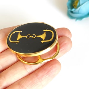 May include: A gold-toned metal ring with a black, oval-shaped top. The top features a gold-colored equestrian bit design. The ring is held in a hand against a white background, with a blue flower in the background.