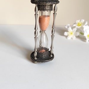 May include: An hourglass with a clear glass bulb containing peach-colored sand. The frame is made of metallic silver-toned material with four vertical pillars, featuring intricate filigree detailing on the top and bottom circular bases.