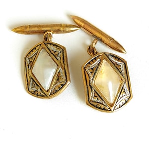 Peut inclure: Une paire de boutons de manchette dorés présentant un cadre hexagonal allongé avec des détails gravés complexes. Chaque bouton de manchette centre une incrustation en nacre irisée en forme de losange et une attache à barre.