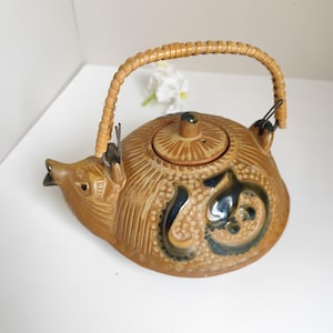 Può includere: Una teiera in ceramica color marrone chiaro a forma di riccio con corpo testurizzato, un piccolo beccuccio e un coperchio rimovibile. La teiera presenta un simbolo smaltato blu scuro sul lato e un manico curvo in rattan intrecciato marrone chiaro fissato con anelli metallici.
