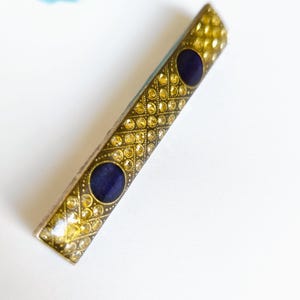 Puede incluir: Una horquilla rectangular dorada con un patrón repetido de pequeños pedrería amarillos. Dos acentos circulares azul oscuro están incrustados en la horquilla. La horquilla es probablemente vintage o antigua.
