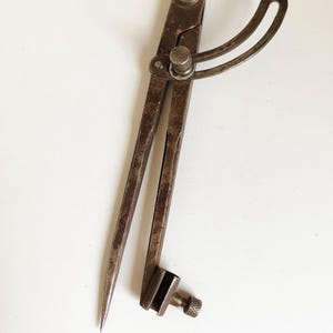 Può includere: Un compasso da disegno in metallo vintage con una patina marrone scuro usurata. Lo strumento presenta due gambe dritte, una con una punta affilata e l'altra con un supporto cilindrico fissato da una vite zigrinata e un braccio di regolazione arcuato.