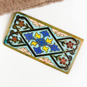 Peut inclure: Pièce décorative rectangulaire en métal avec un fond bleu clair et un motif central en losange noir, bleu et jaune. Les bords sont soulignés d'or, avec des accents floraux rouges et verts. Un tissu marron est partiellement visible en haut à gauche.