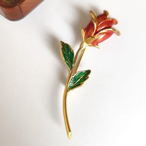 Peut inclure: Une broche en forme de rose en métal doré, avec des pétales en émail rouge et des feuilles en émail vert. La broche représente une seule tige et fleur de rose. La broche est sur une surface blanche.
