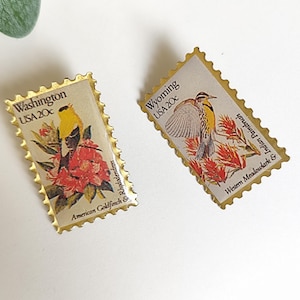 Peut inclure: Deux broches en émail rectangulaires en forme de timbres-poste avec des bords festonnés dorés. La broche de gauche représente un chardonneret jaune sur un rhododendron rouge avec le texte Washington USA 20c. Celle de droite montre une sturnelle et une castilléja avec le texte Wyoming USA 20c.