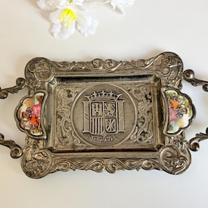 Puede incluir: Bandeja de servir plateada ornamentada con asas y bordes decorativos. Presenta un emblema central con una corona y un escudo, y pequeños insertos de porcelana coloridos. La palabra "ESPANA" es visible.