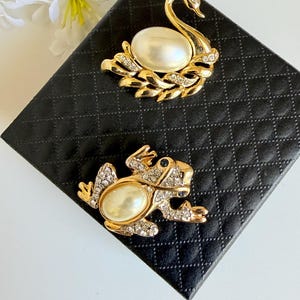 Peut inclure: Deux broches décoratives. L'une est un cygne doré avec un corps en perle et des accents de cristal. L'autre est une grenouille dorée avec un corps en perle et des accents de cristal. Les deux broches sont sur une surface noire matelassée.