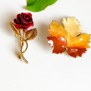 Peut inclure: Deux broches vintage : une rose rouge et une feuille. La broche en forme de rose présente une rose en émail rouge détaillée avec des feuilles et une tige dorées. La broche en forme de feuille a un dégradé d'émail orange, jaune et marron. Une feuille verte est en arrière-plan.