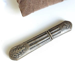 Puede incluir: Un estuche para agujas plateado con un diseño floral ornamentado. El estuche rectangular tiene un cuerpo acanalado y extremos decorativos. Se encuentra sobre una superficie blanca, con un elemento de tela marrón en el fondo.