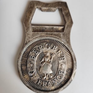 May include: Vintage silver-toned bottle opener with a circular emblem featuring a woman holding a staff, surrounded by the text "DE BERGER FRANCE". The opener has a classic design with a bottle cap removal slot.
