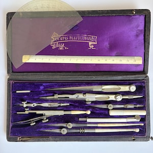 May include: A vintage drafting instrument set in a black case with a purple velvet interior. The set includes various metal compasses, a ruler, and a protractor. The case features the text COMPAS PERFECTIONNES printed in gold on the fabric.